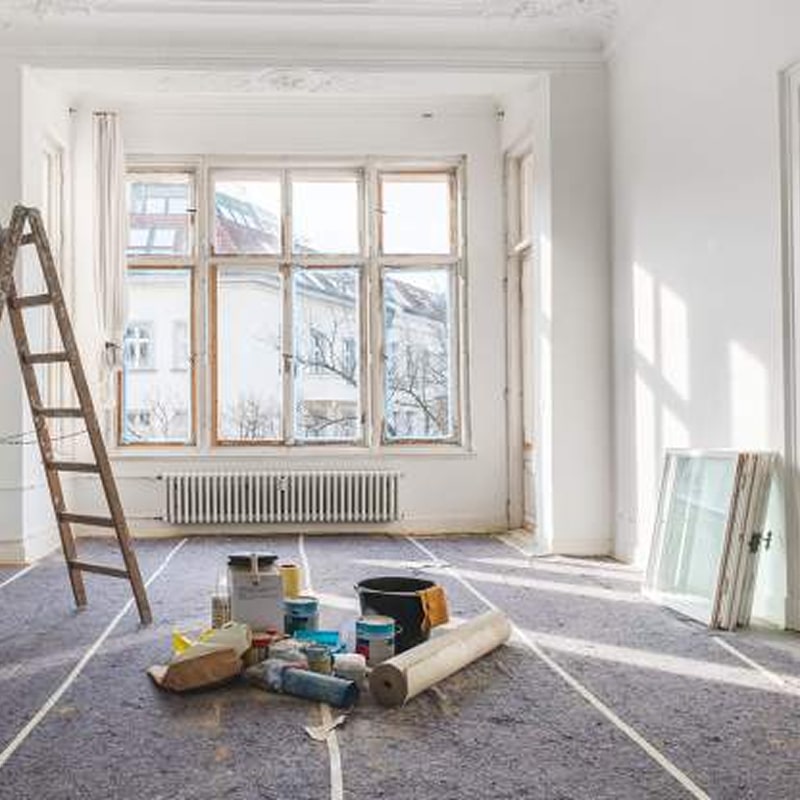 Chambre en rénovation avec échelle en bois, fournitures de peinture, rouleaux et sol protégé. Lumière du soleil par la grande fenêtre.