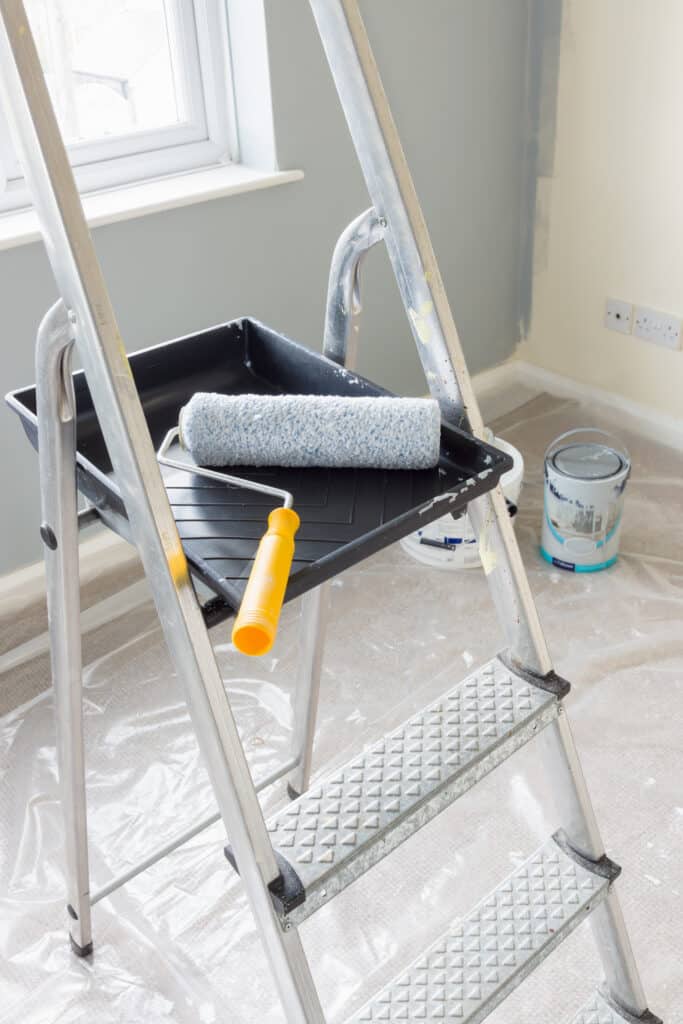 Painting and decorating with a roller and tray on a set of metal step ladders with cans of paint and a dust sheet in the background